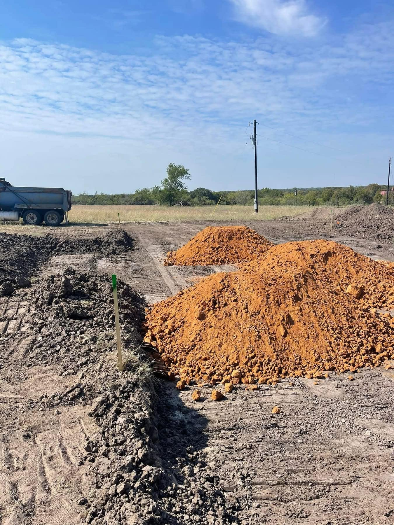 Piles of orange gravel at a rural construction site under a blue sky, with a truck and utility pole in the background