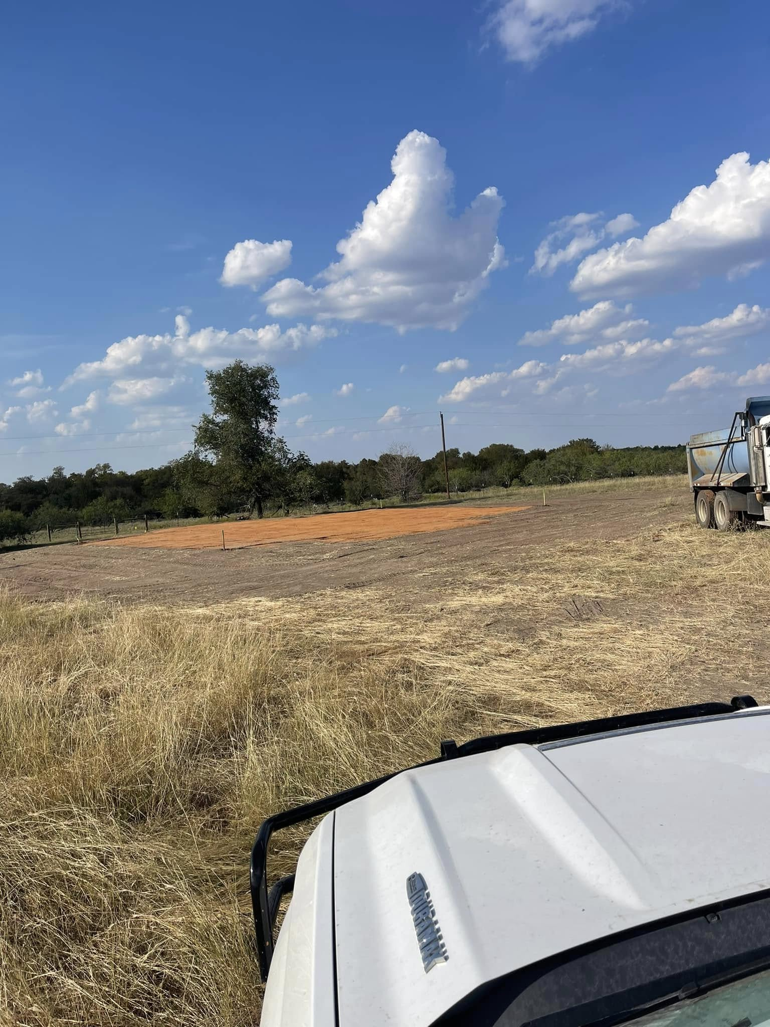 Dry field under a blue sky with clouds, seen from a vehicle, with a truck at the right edge.