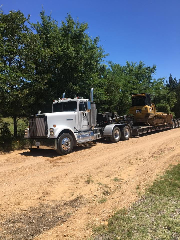 White semi truck towing a yellow bulldozer on a dirt road beside trees under a blue sky