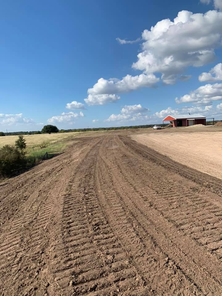 Rural dirt road under a blue sky with clouds, leading past fields to a distant barn and house