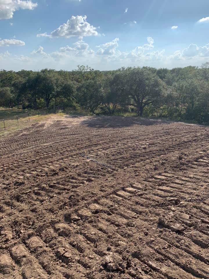 Freshly plowed dirt field with tire tracks, bordered by trees under a blue, partly cloudy sky