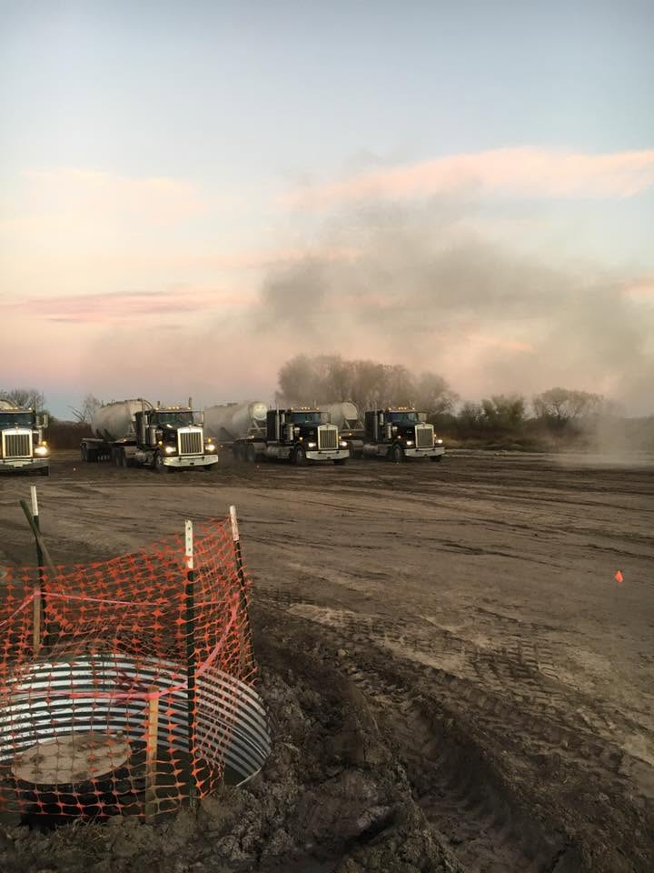 Construction trucks on a dusty dirt lot at dusk, with smoke or dust rising in the background.