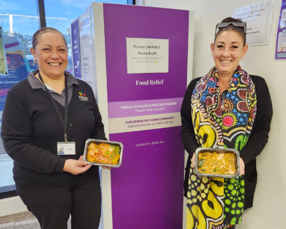 Two people holding meal trays beside a purple Food Bank donation