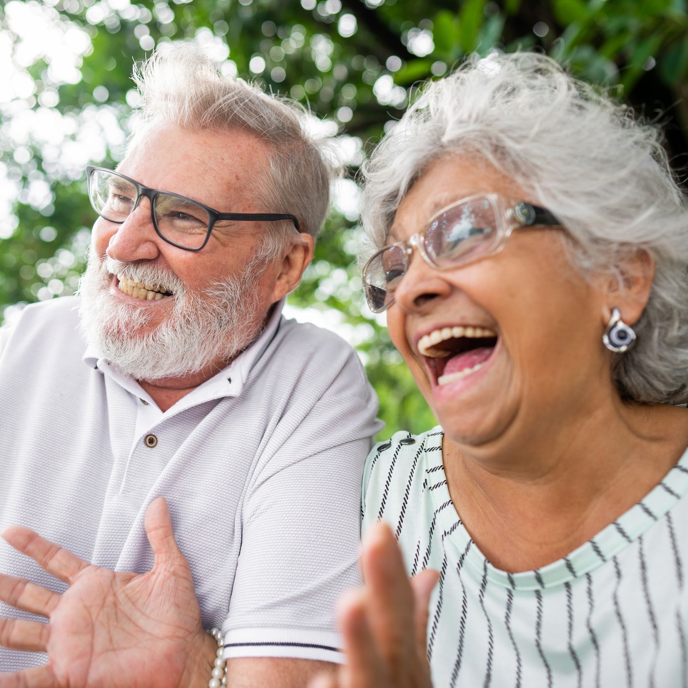 Elderly couple laughing outside, wearing glasses. Man has a white beard. Woman has silver hair.