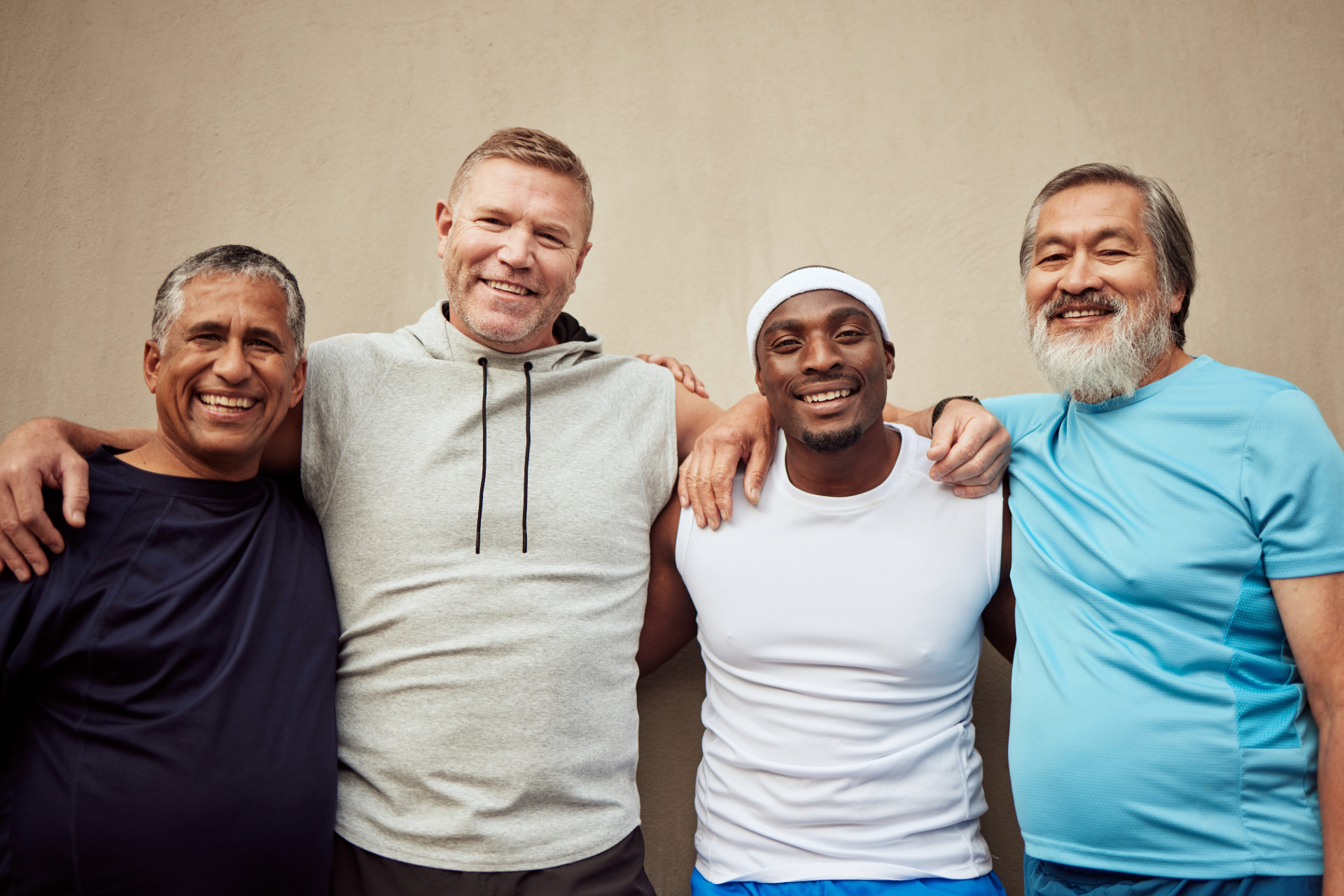 Four smiling friends standing close together against a plain background, wearing casual shirts.