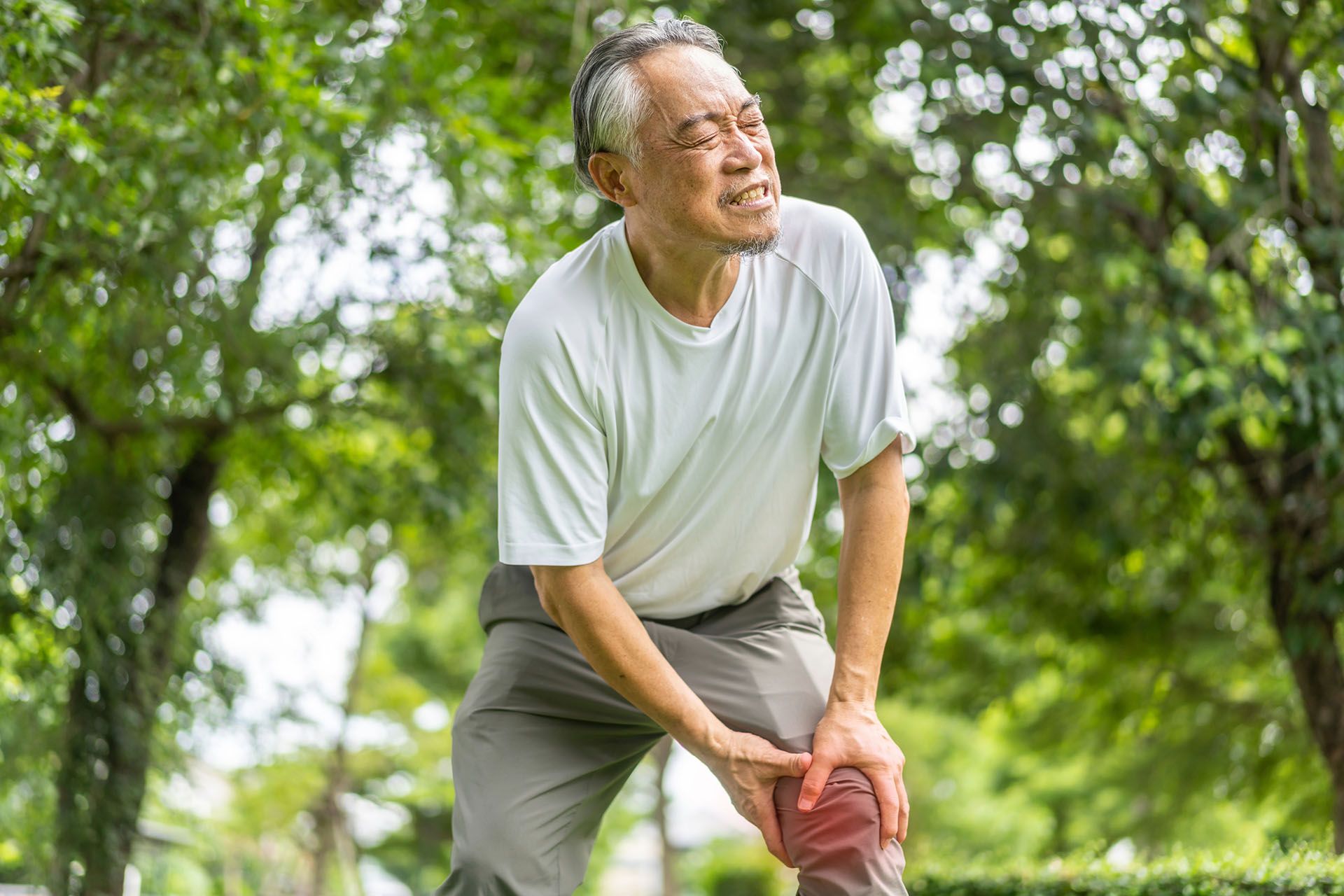 An elderly man is kneeling down in a park holding his knee in pain.
