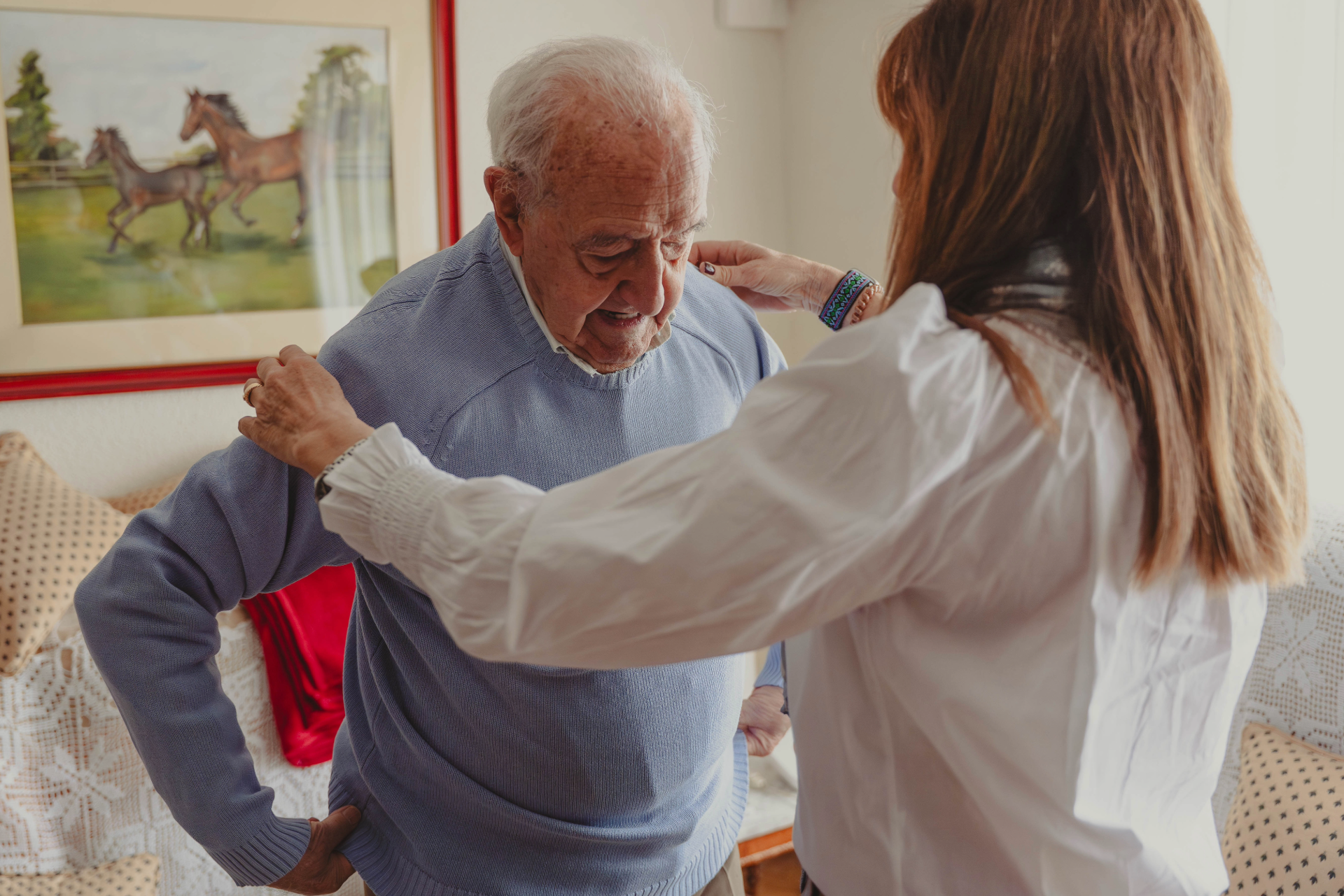 Woman helping an elderly man put on a sweater in a cozy living room.