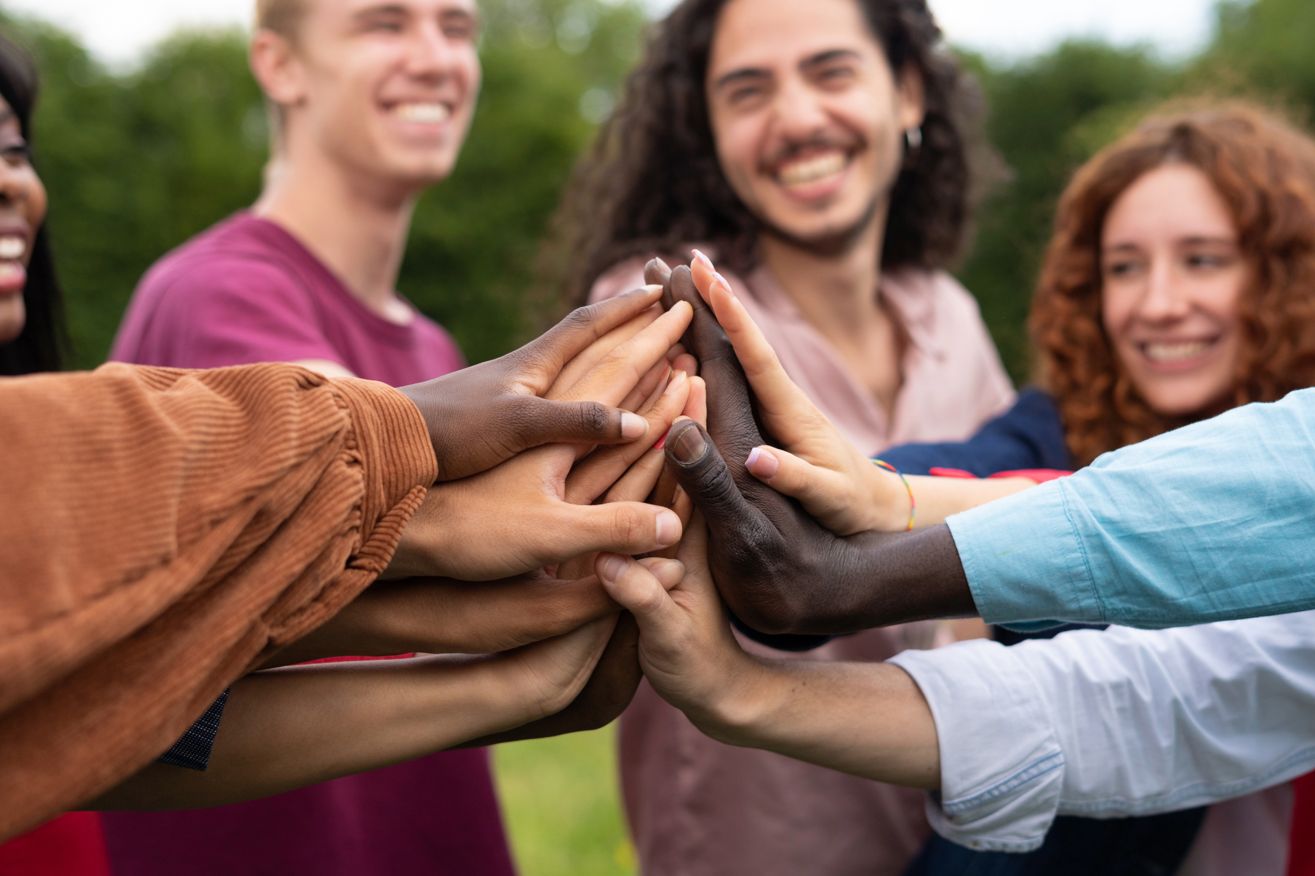 Hands stacked together in a group hug outdoors, with smiling people in the background.