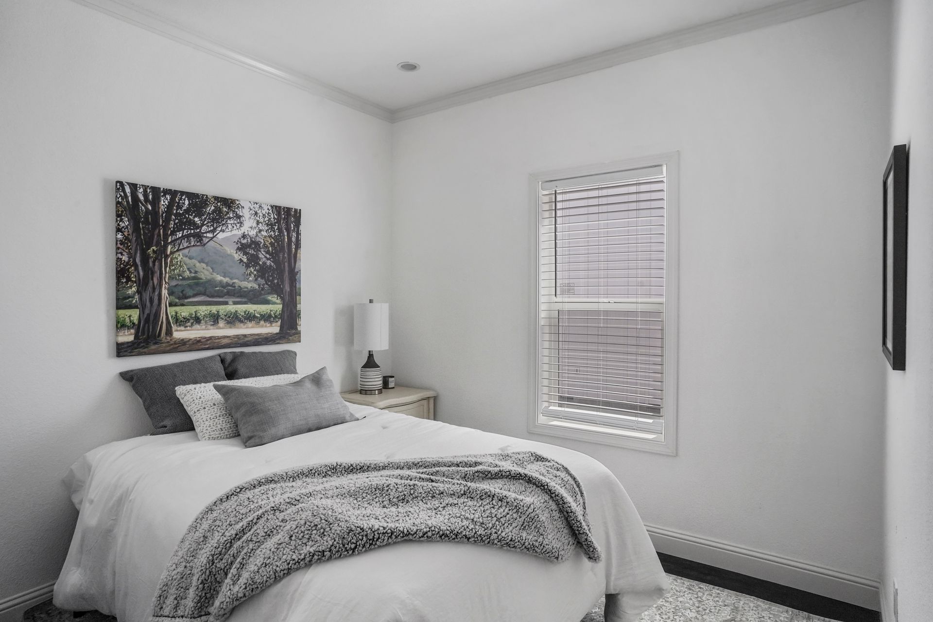 Perpetual Homes ADU: Bedroom with white walls, bed with pillows and blanket, bedside table, window with blinds.