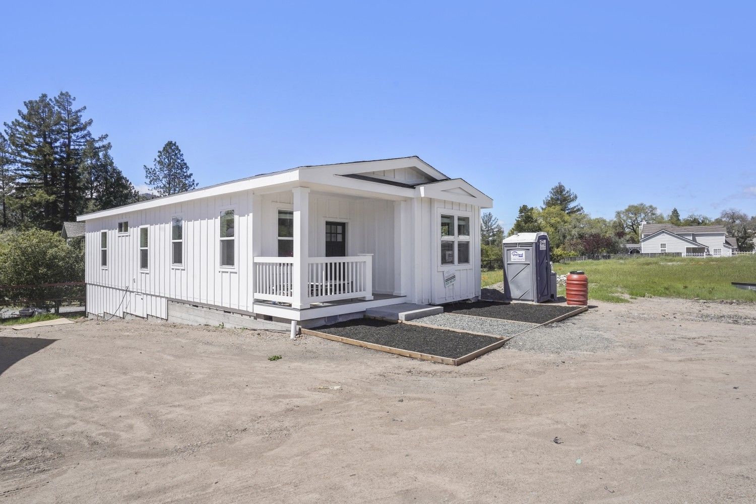 Perpetual Homes ADU: White tiny house with a porch and outhouse in a gravel lot under a blue sky.