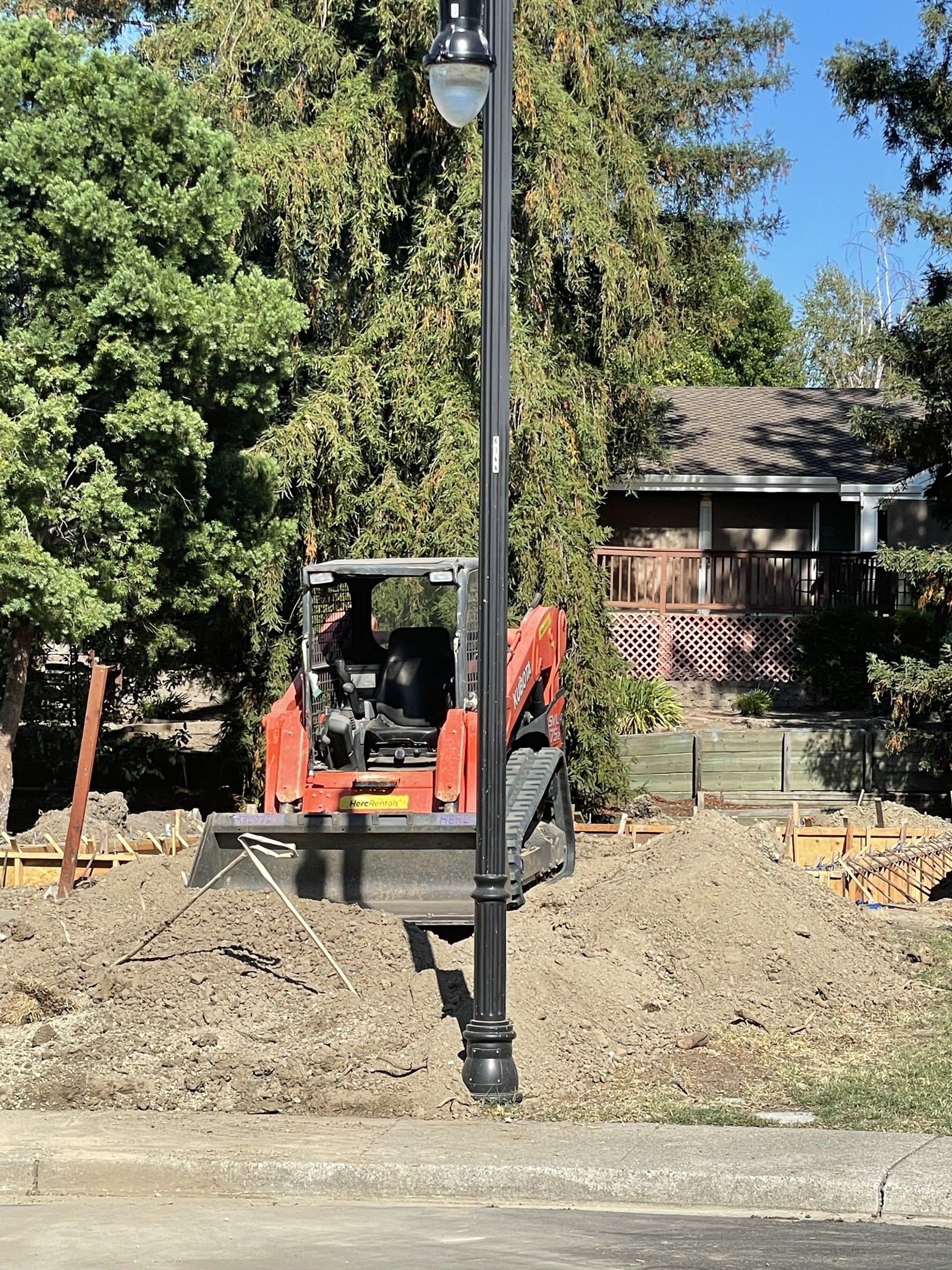 Construction site: Mini excavator operator digging near a lamppost and small house.