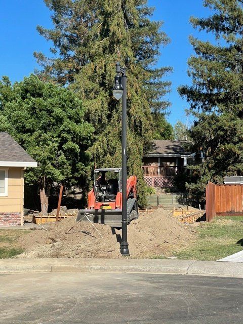 Construction site with mini excavator, dirt pile, and lamppost on a sunny day. Houses and trees in the background.
