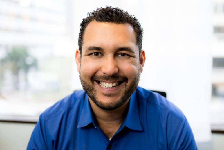 Smiling man in blue shirt, light-skinned with dark hair and beard, indoors with a bright background.