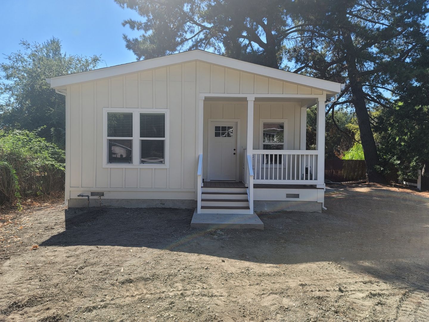 Perpetual Homes ADU: Small, light-colored house with porch, steps, and door. Windows on the left, trees in background.