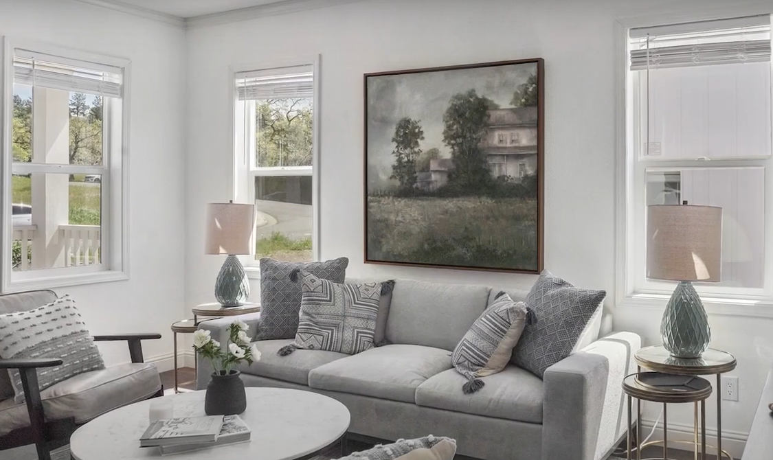 Perpetual Homes ADU: Living room with gray sofa, landscape painting, two windows, and two table lamps.