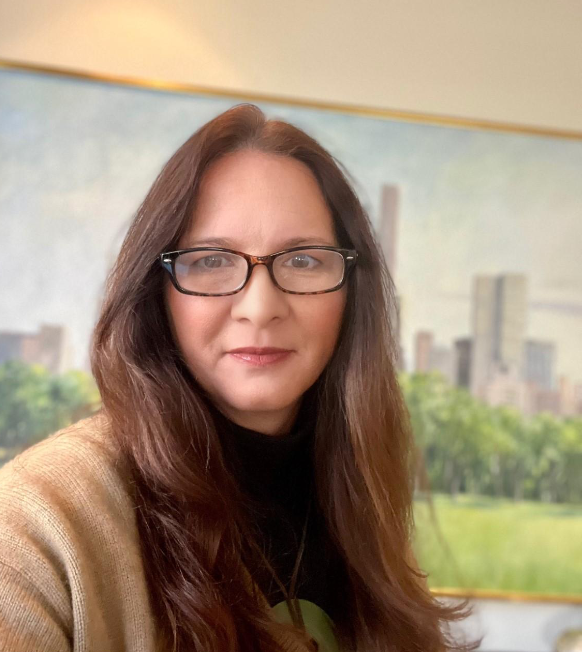 Woman with glasses smiles; brown hair, black turtleneck, beige cardigan, background painting of a city.