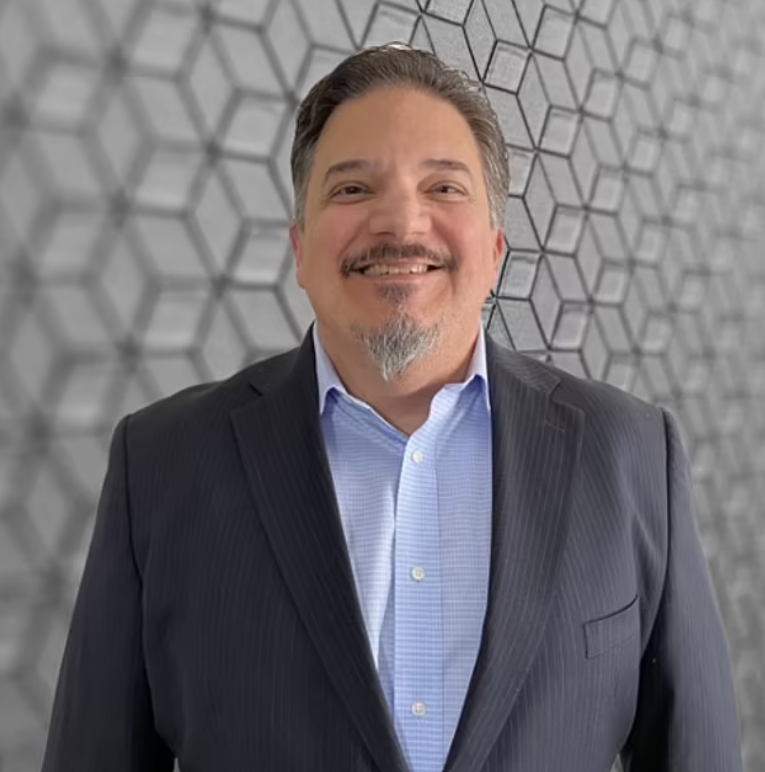 Man in a blue shirt and suit jacket smiling in front of a geometric wall.