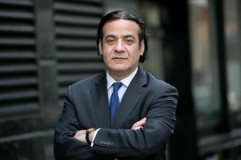 Man in a dark suit with arms crossed, standing outdoors in front of a building.