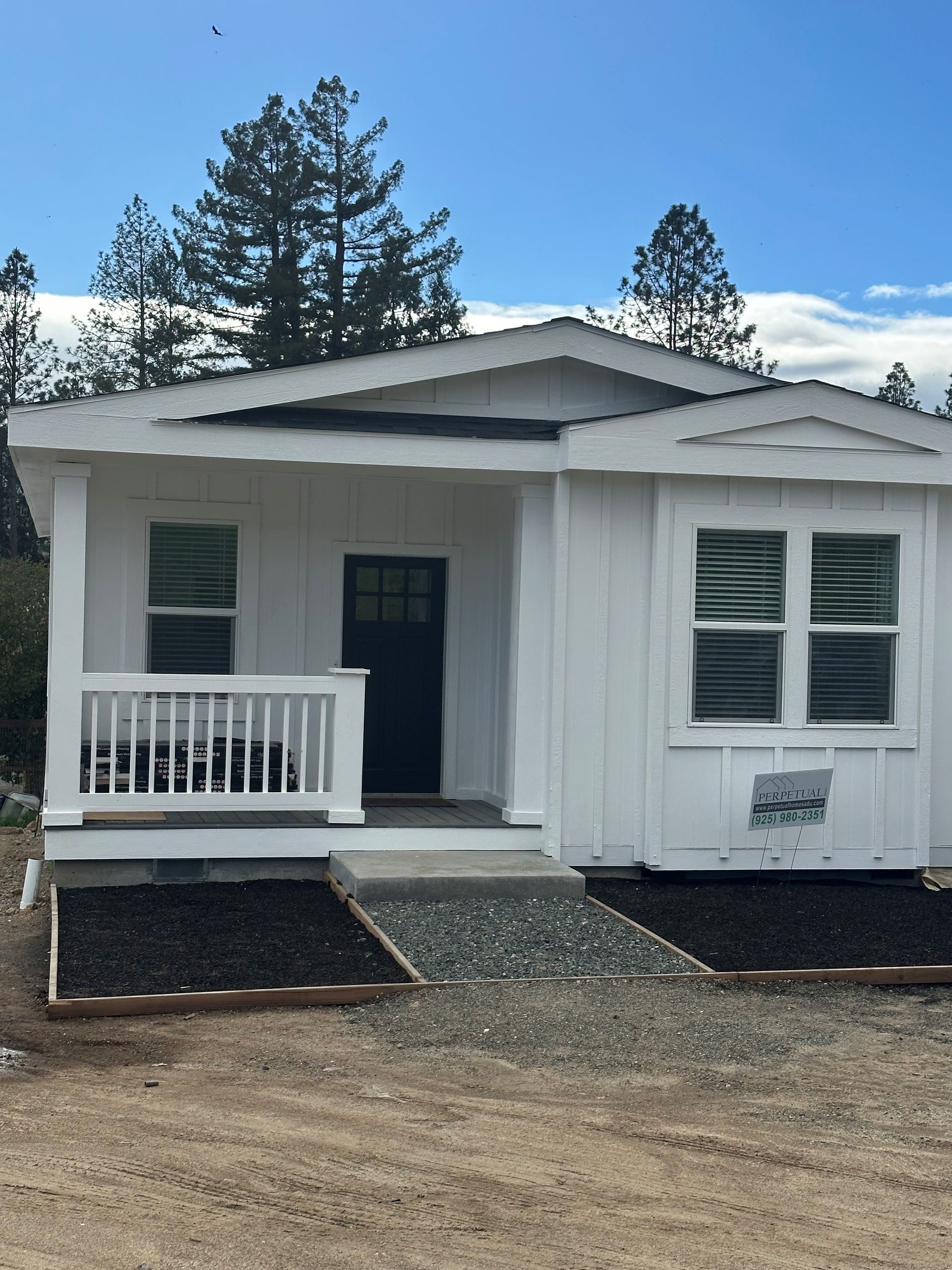 Perpetual Homes ADU: White cottage with a porch, dark door, and windows, set against a backdrop of trees and a blue sky.