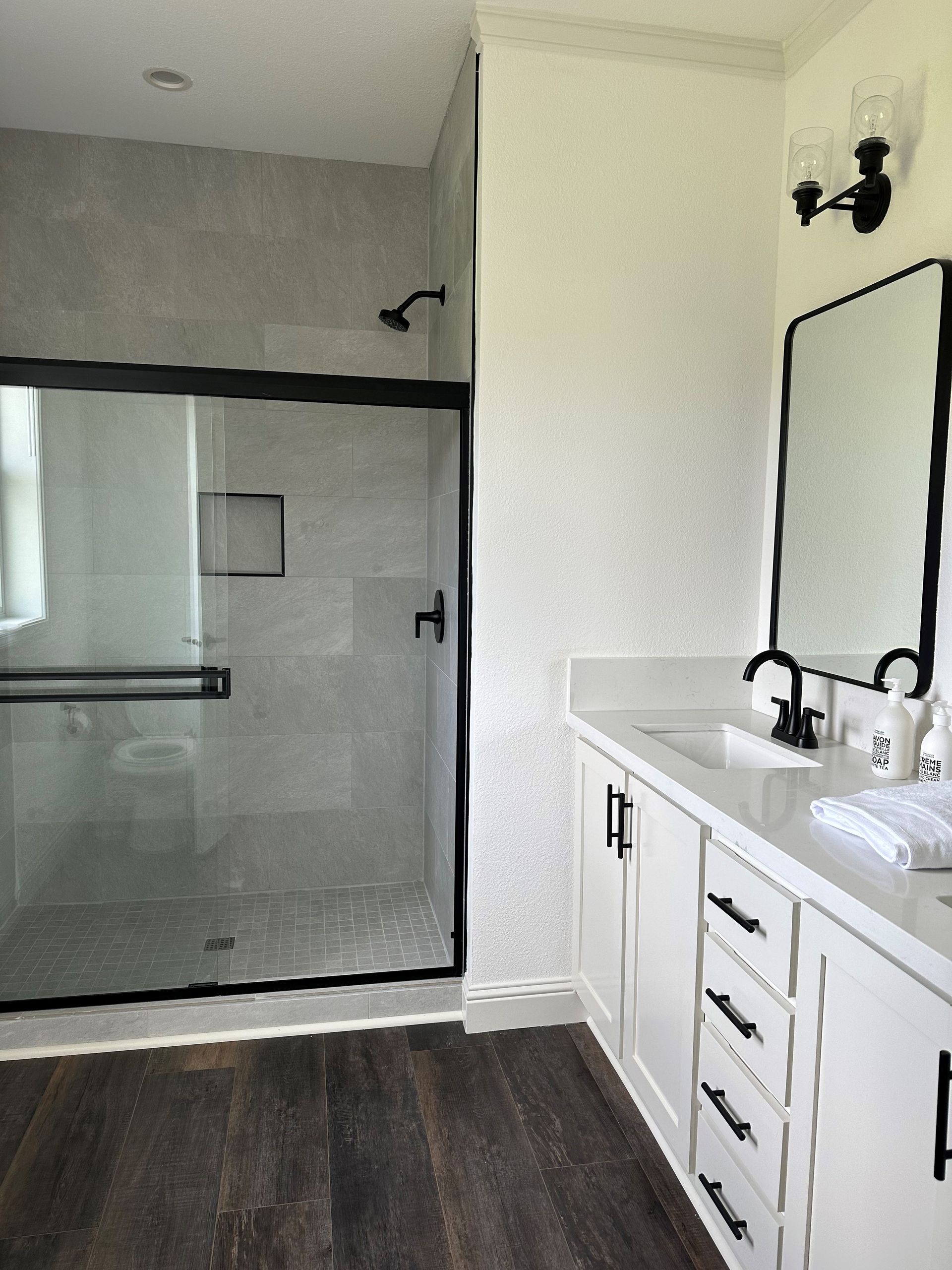 Perpetual Homes ADU: Bathroom with glass shower, white vanity, black accents, and dark wood floor.