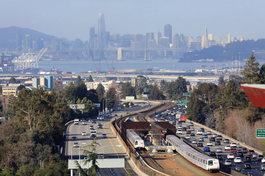 Highway traffic and train alongside a station, with San Francisco skyline and bridge in the distance.