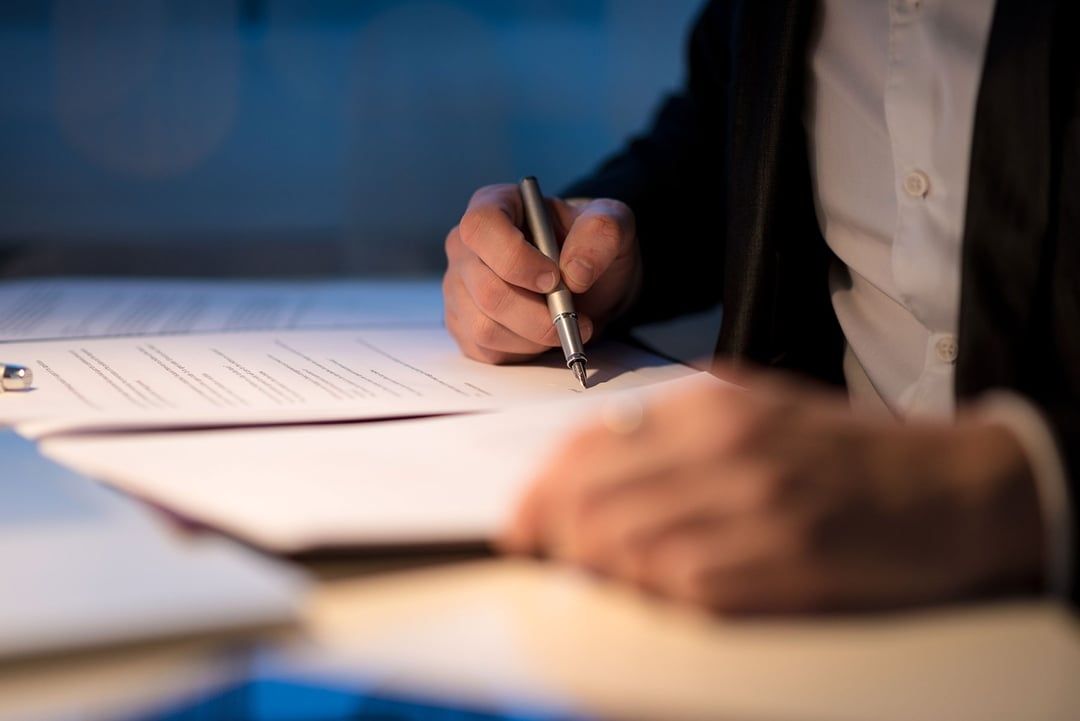 A person in a business suit signs a legal document with a pen at a desk under soft lighting.