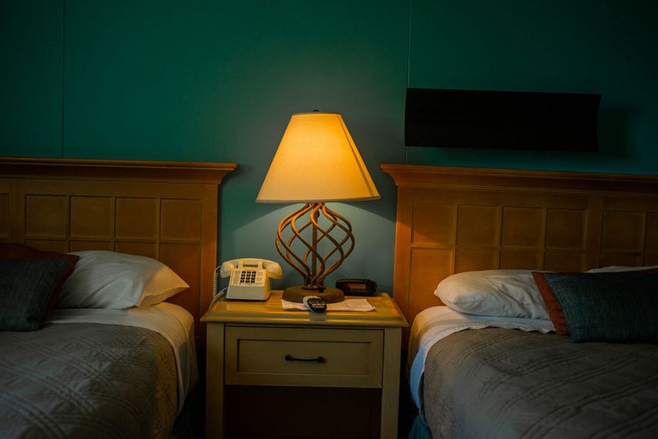 Two beds in a dim motel room with a lit lamp on a bedside table between them