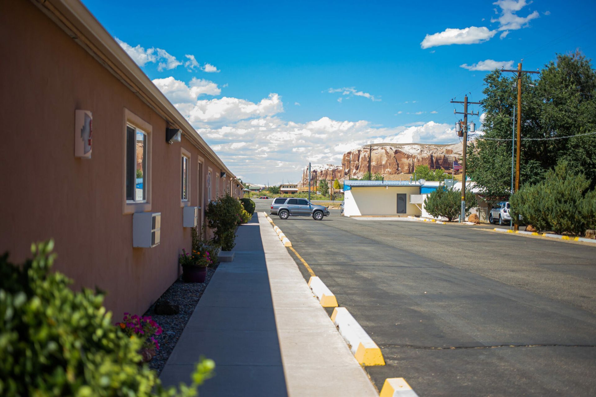 Quiet street beside tan building under a blue sky, with parked cars and distant trees.