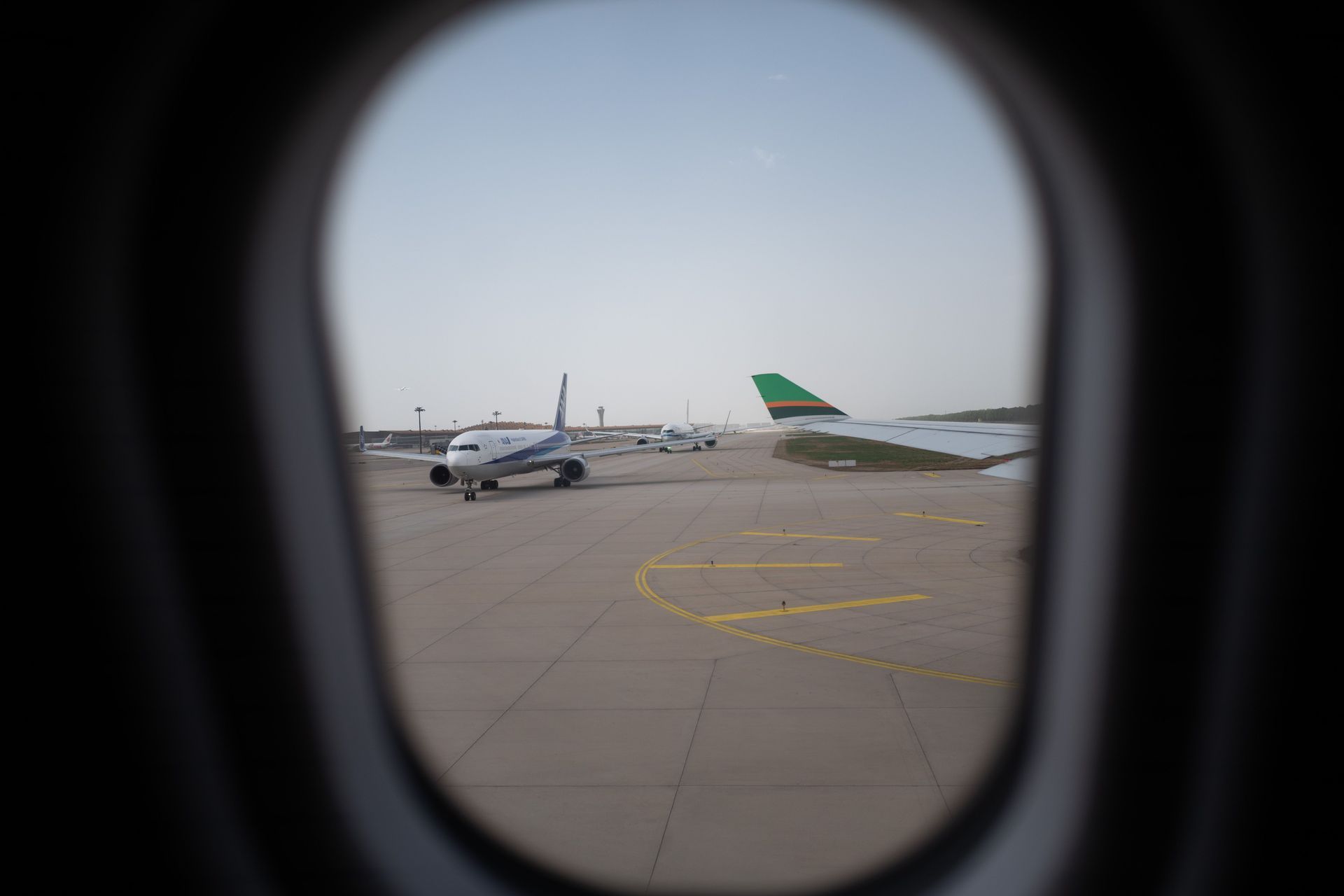 View through an airplane window of a runway with parked planes, including one with a green tail.
