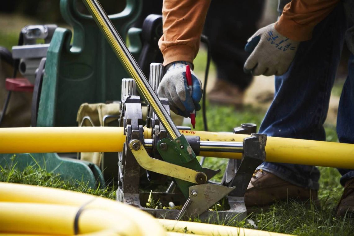 Worker in blue jeans cutting yellow pipe with a tool, outdoors on grass.