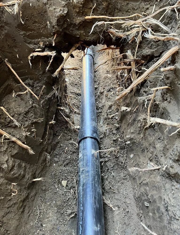Black pipe running through a dirt trench with exposed roots and soil around it