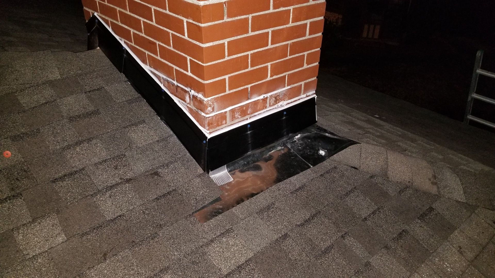 A cat is laying under a brick chimney on a roof.