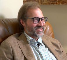 Smiling man with glasses and beard sitting in a brown leather chair, wearing a tan blazer.