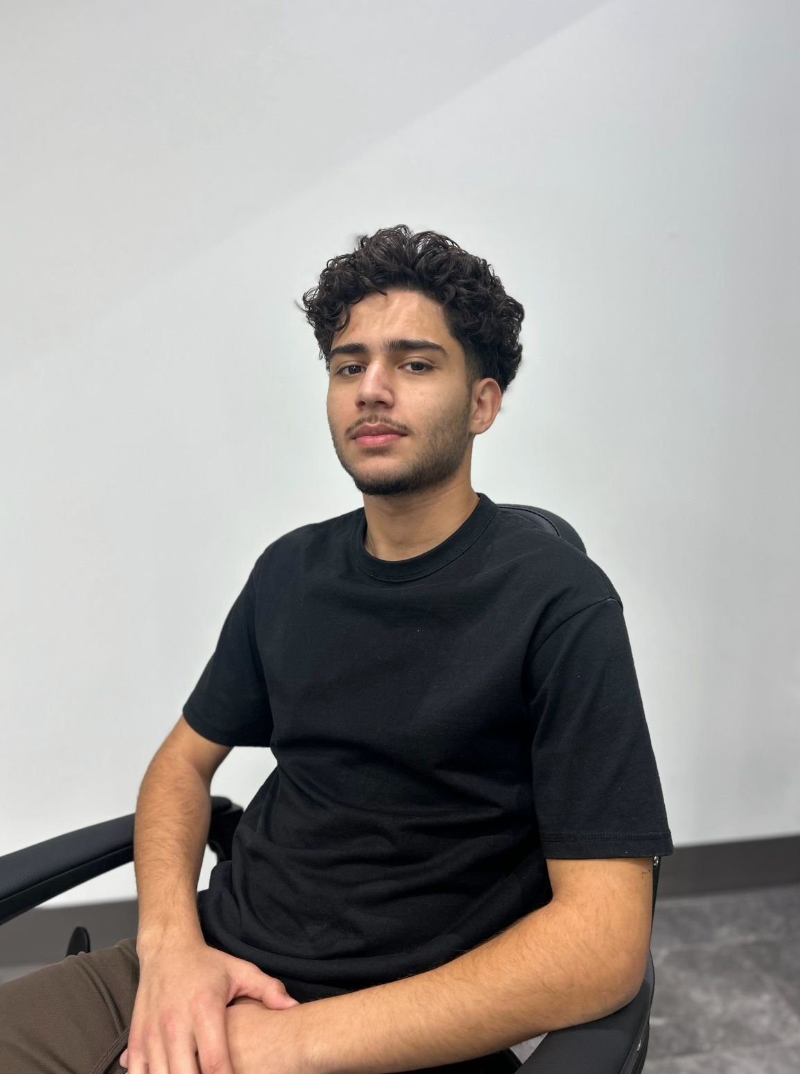 A person with curly hair wearing a black t-shirt sits in a chair in front of a white wall.