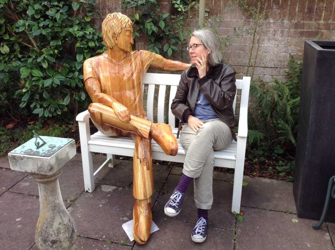 A human lady sitting on a bench with a sculpture of a man made by Ingrained Culture