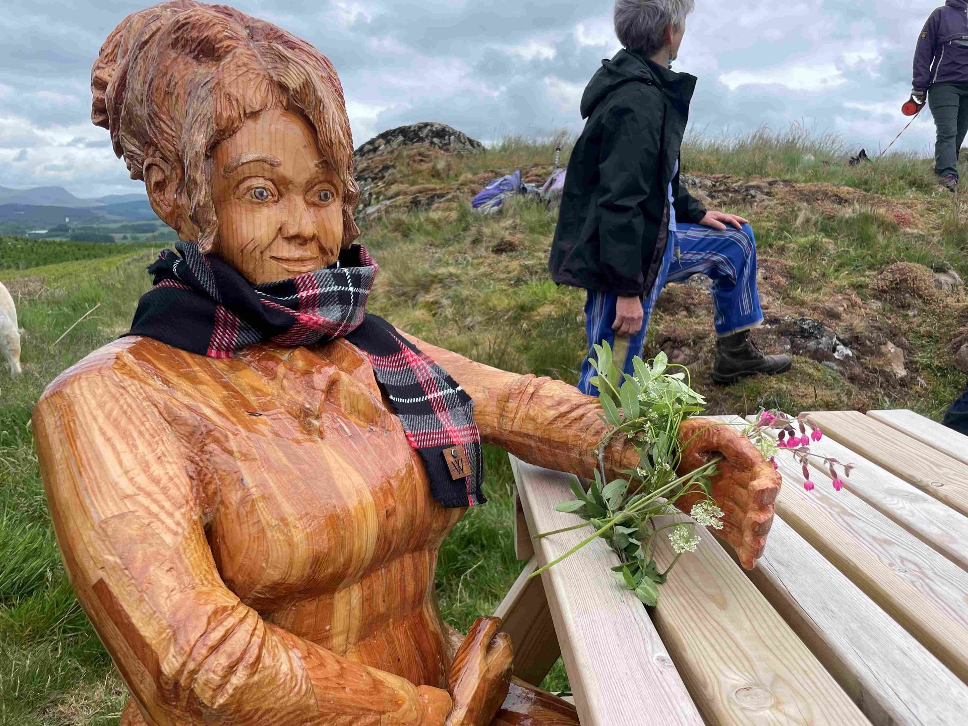 Hand-carved wooden sculpture, The Storyteller, seated on a bench and wearing a witches tartan scarf.