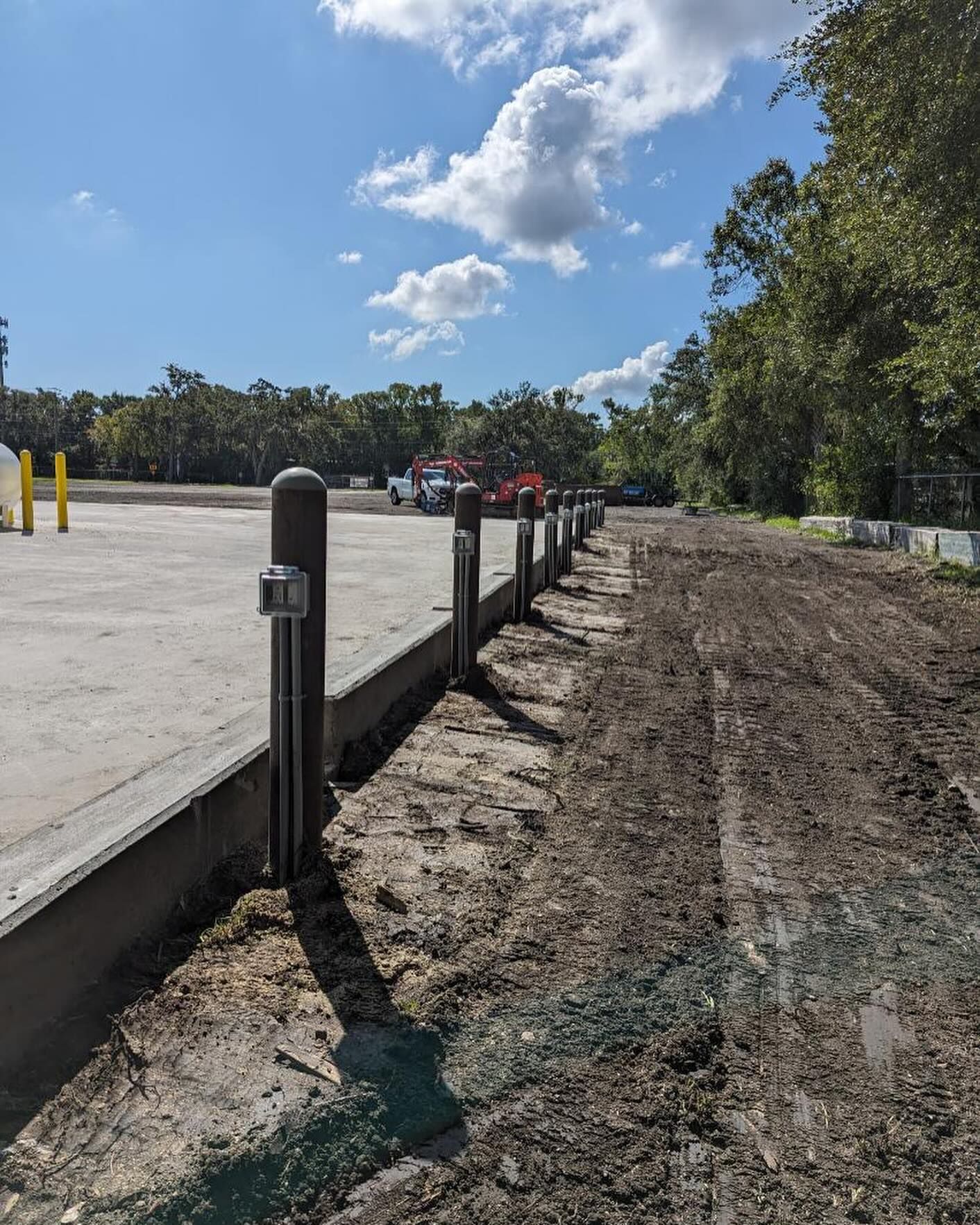 Concrete parking lot beside a dirt path, with bollards, trees, and a blue sky with clouds.