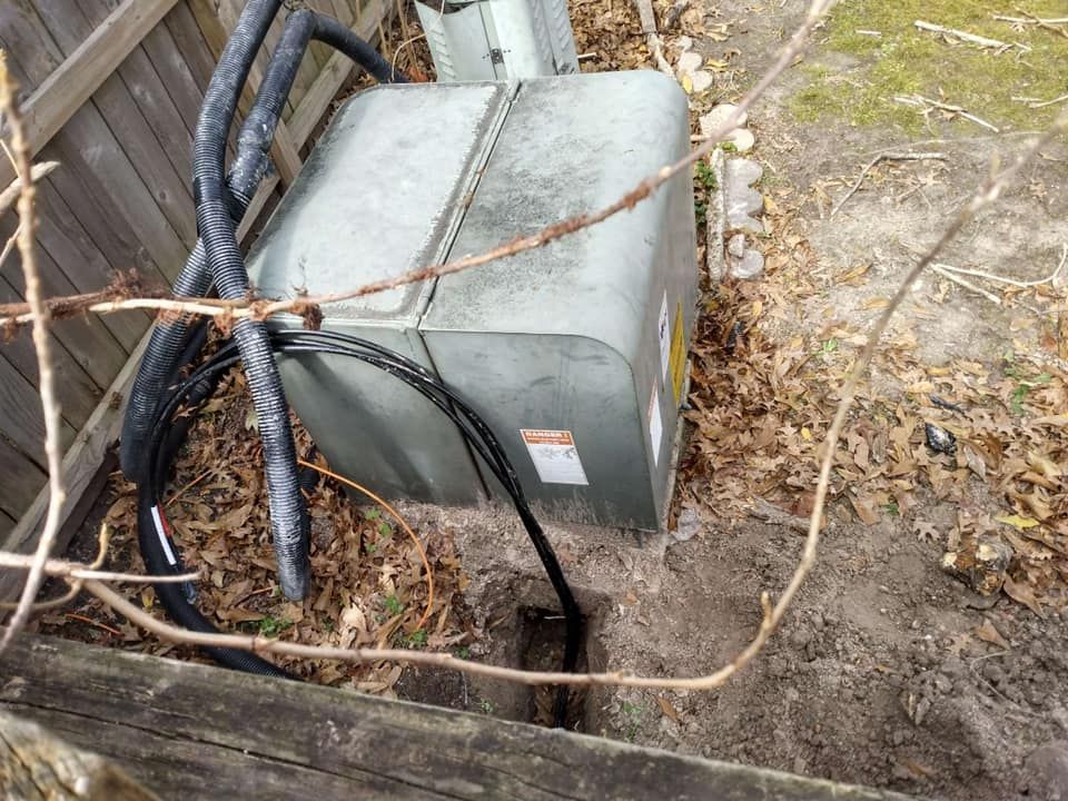 Gray electrical box beside pipes and cables in a dirt trench, near a fence and dry leaves