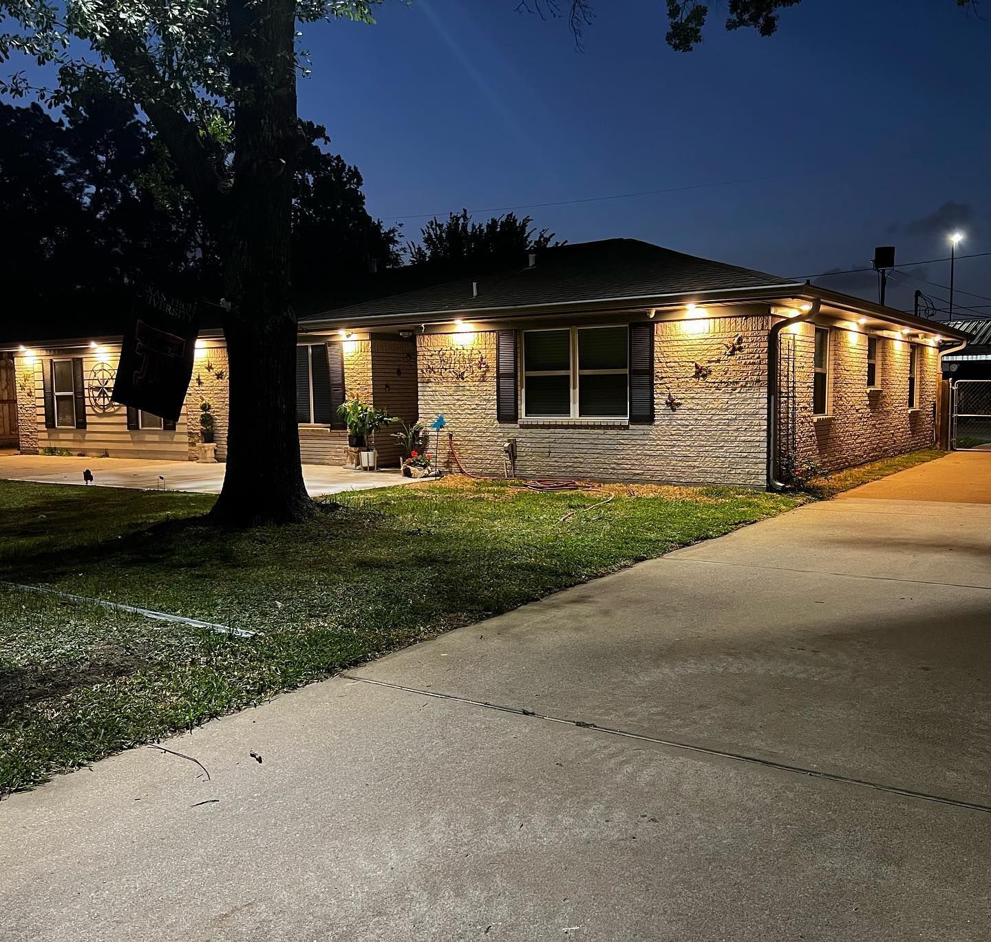 Brick house at dusk with warm porch lights, a large tree, and a driveway in front