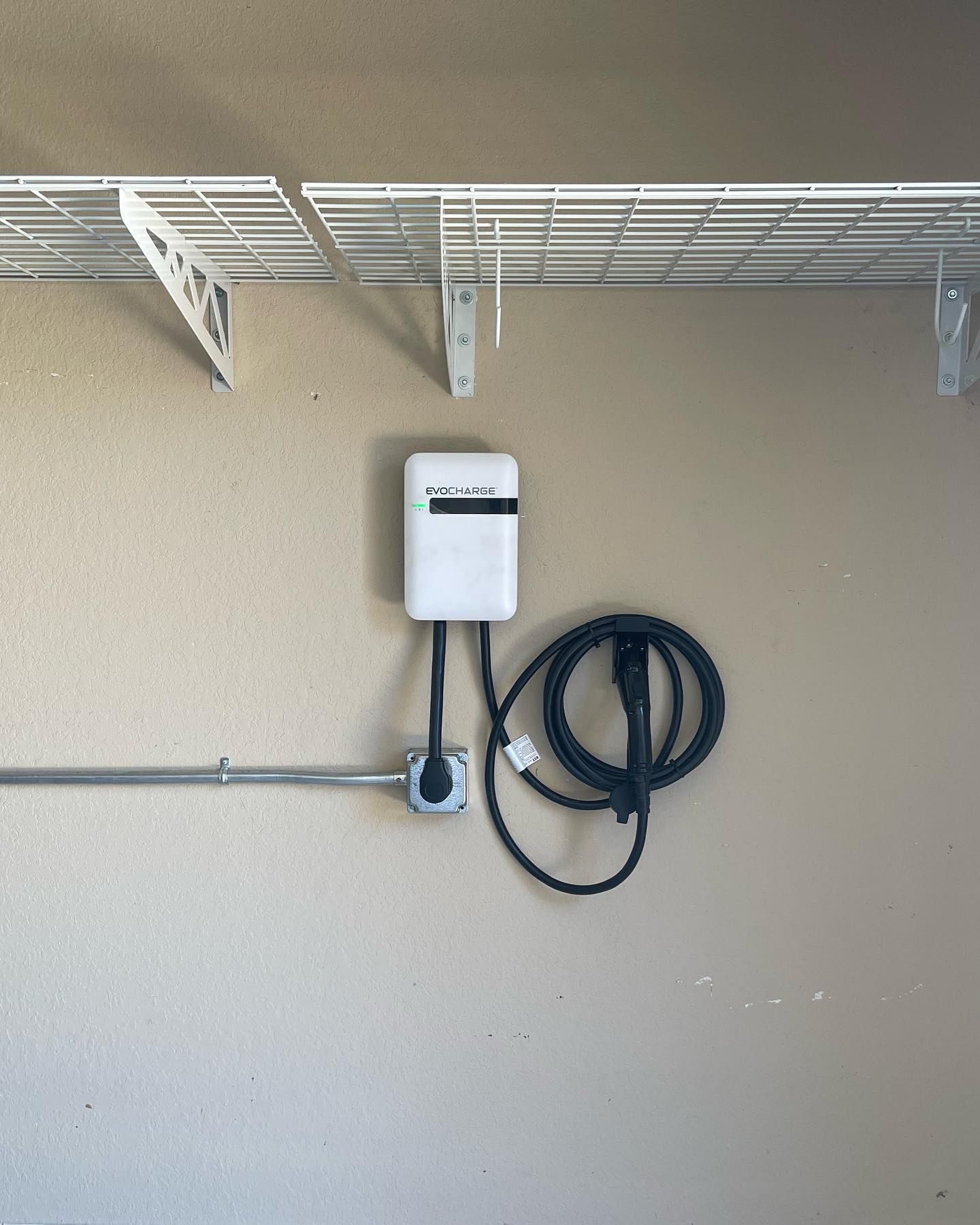 Garage wall with white electrical box, conduit, and coiled black cable beneath wire shelving.
