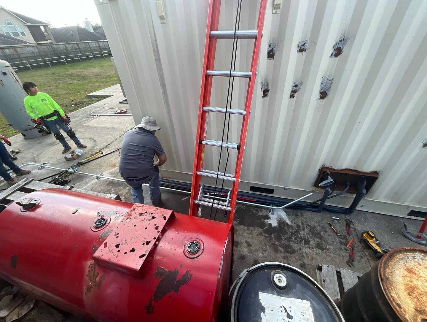 Workers beside a red tank and ladder against a white corrugated wall outdoors