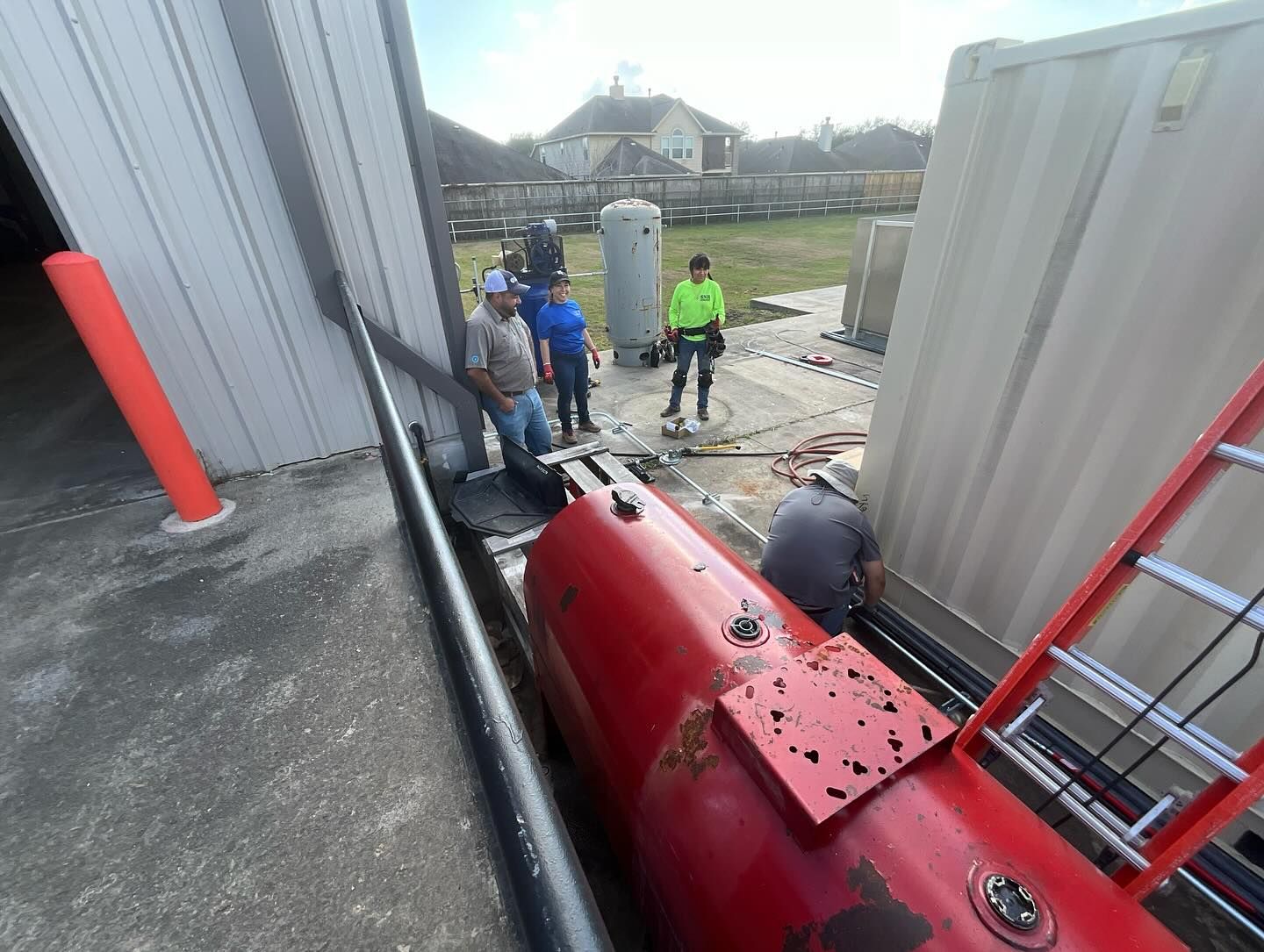 Workers inspect a red industrial machine in a narrow alley beside metal buildings.