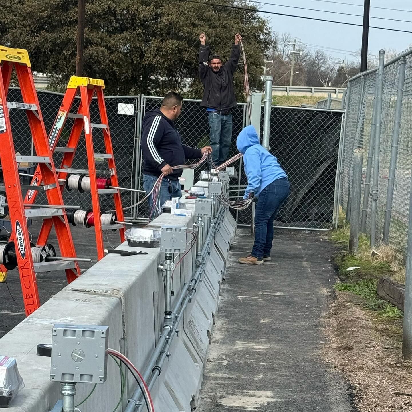 Workers installing gray equipment boxes along a fenced outdoor path beside orange ladders