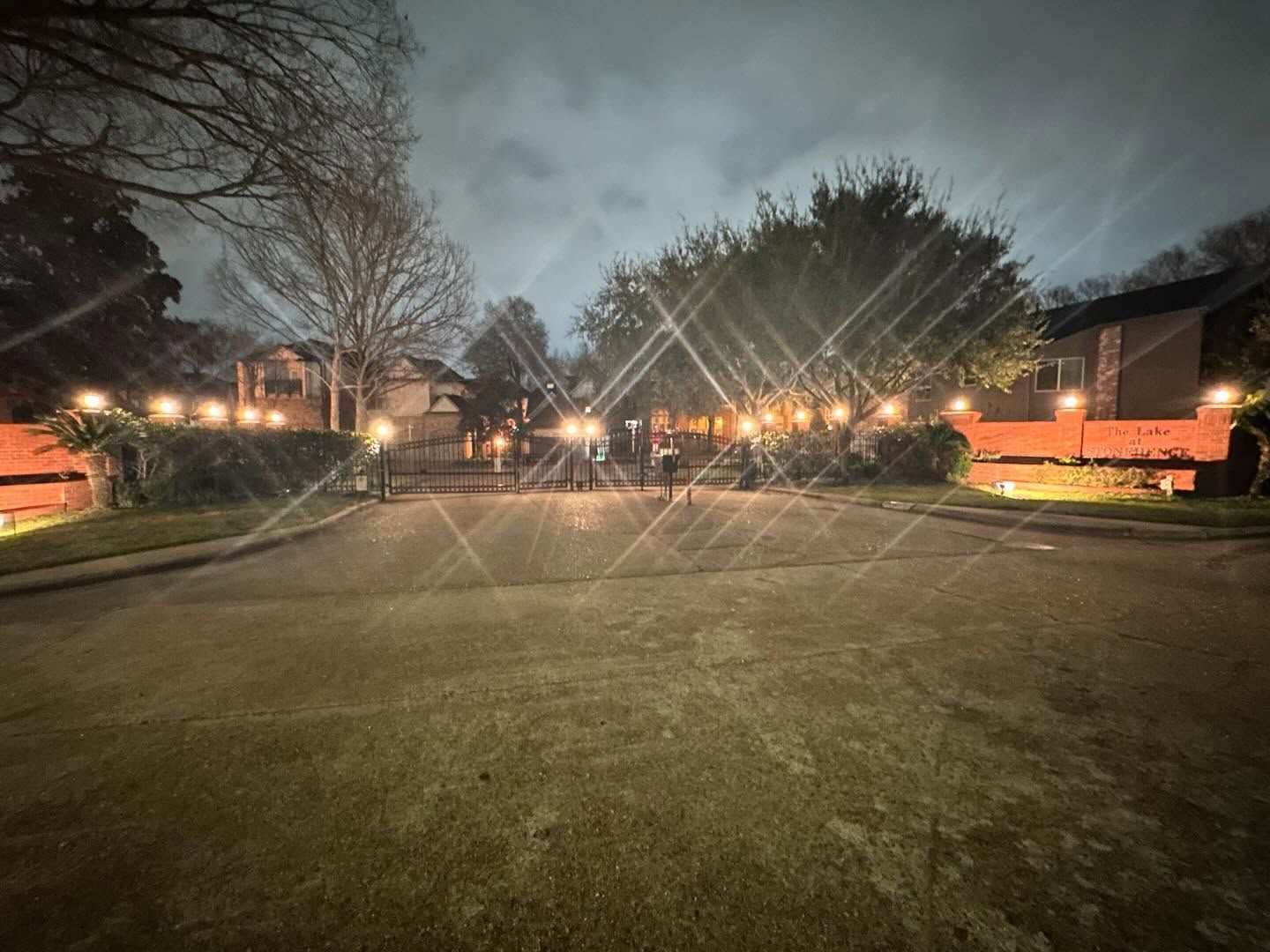 Lit tennis courts at dusk with netting, trees, and buildings in the background