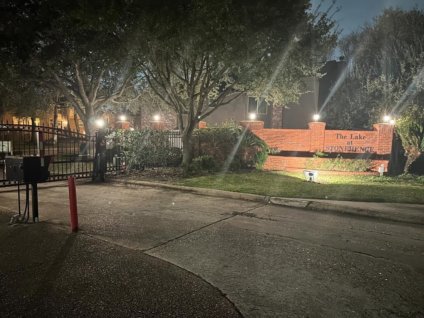 Nighttime view of a lit brick entrance sign beside a tree-lined road and fence