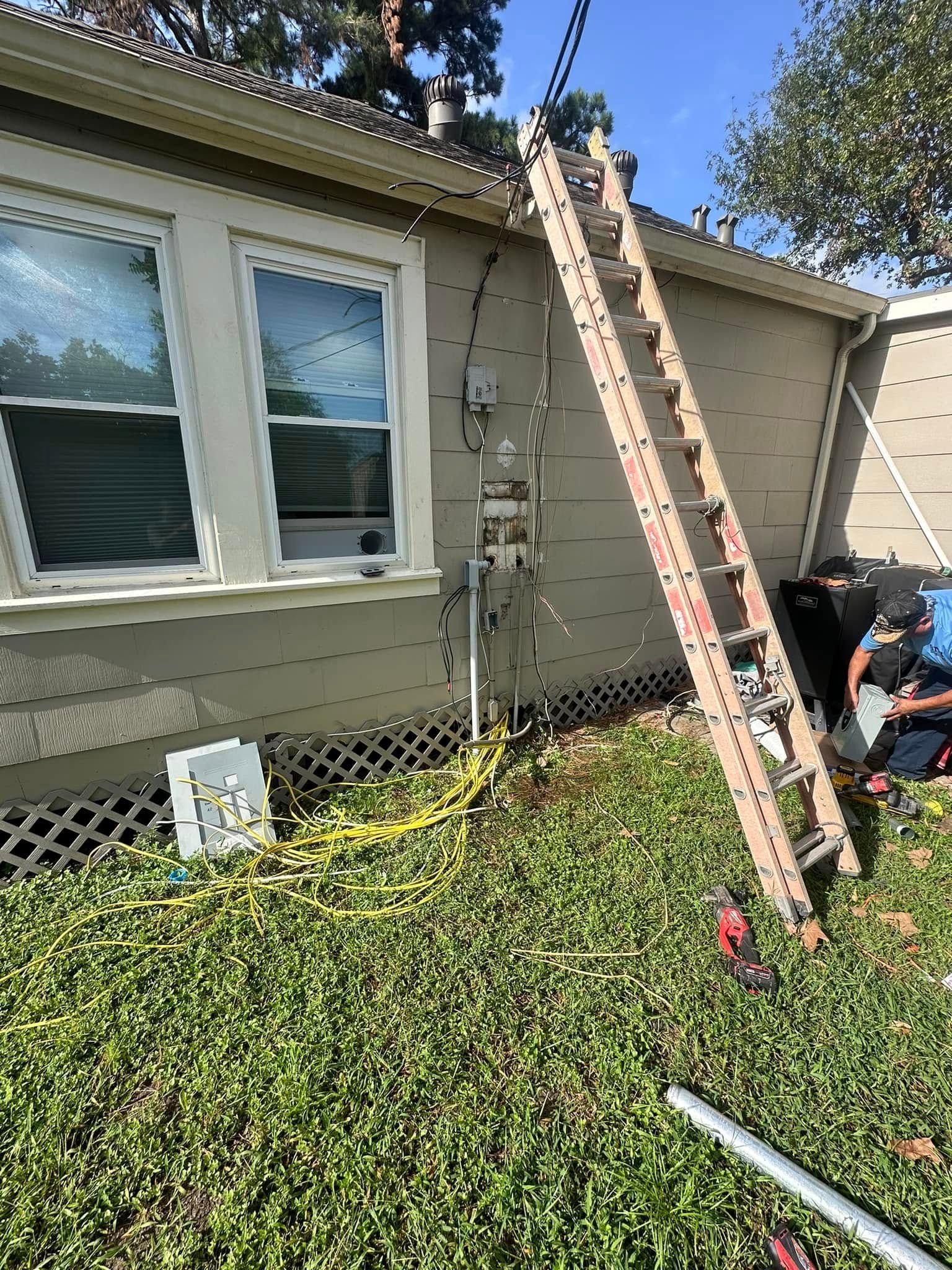Ladder leaned against a house wall beneath windows, with tools and a worker partly visible in the yard.