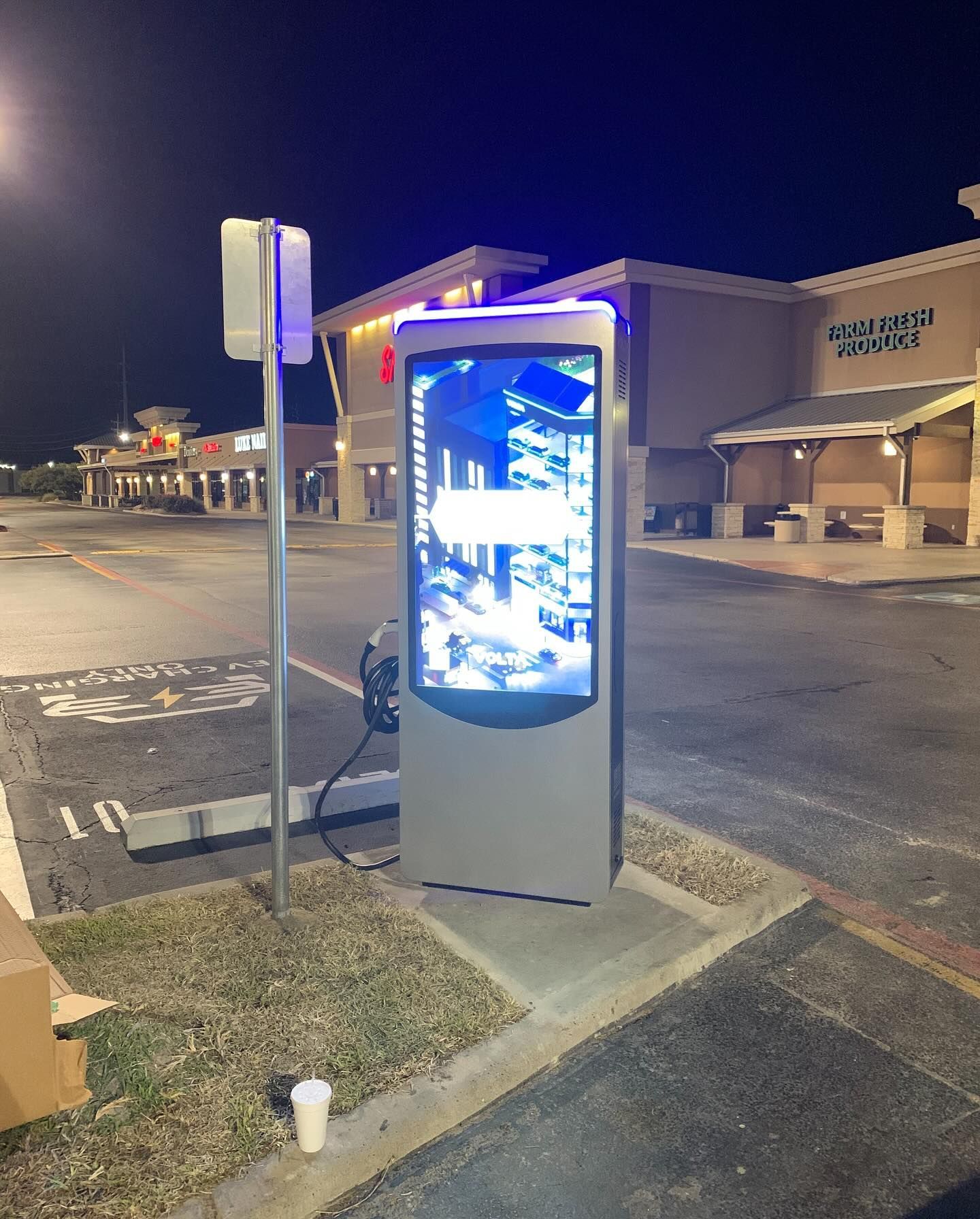 Nighttime parking lot with a glowing digital kiosk beside storefronts and parked cars.