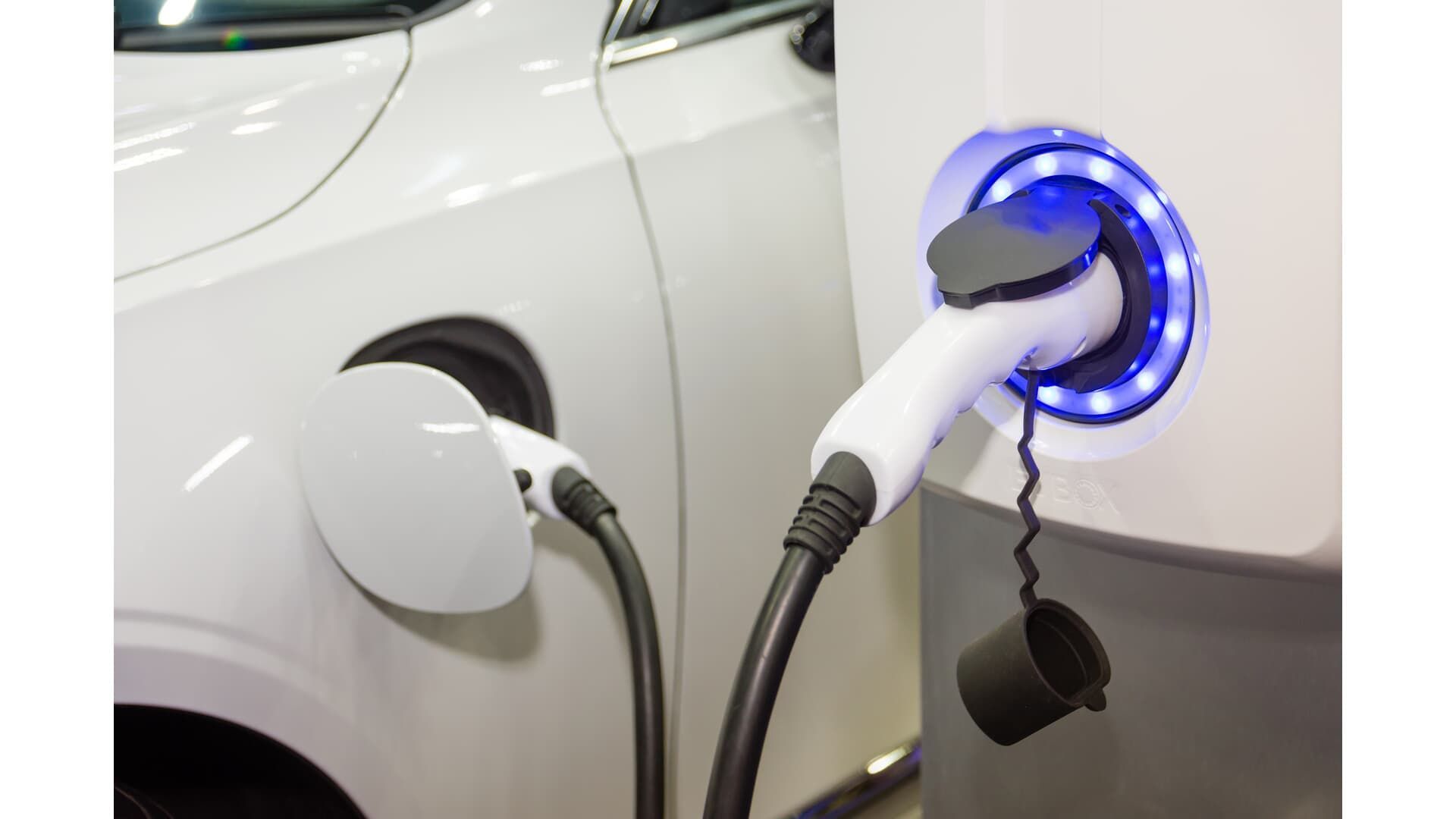 Electric car charging at a public station with a blue-lit connector plugged into a white vehicle