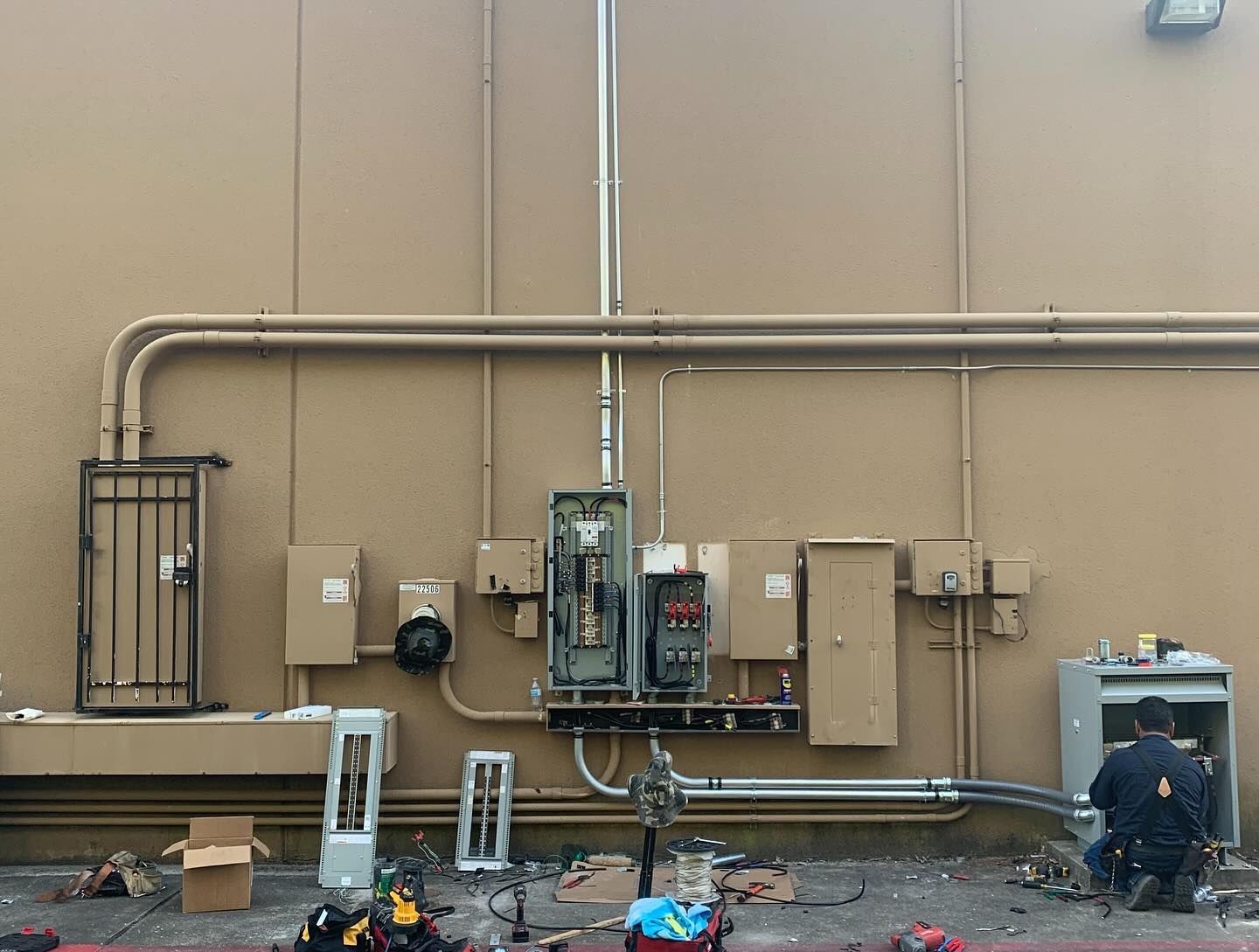 Outdoor utility wall with electrical panels, pipes, and scattered equipment; worker crouches at right.