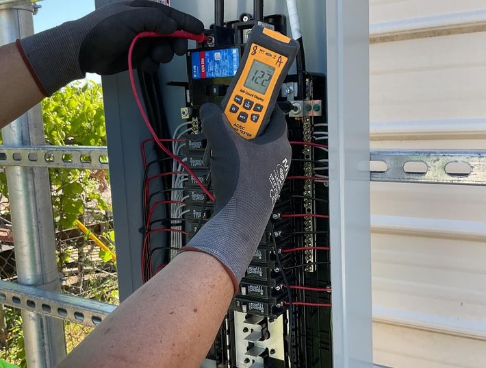 Gloved worker testing electrical wiring in an open control panel with a digital multimeter outdoors